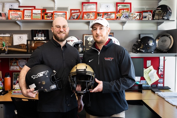 MIT alumnus Kodiak Brush and MIT Football Head Coach Brian Bubna hold two football helmets. Behind them are shelves holding sports photos and awards.