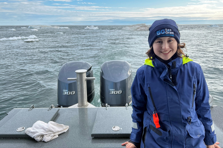 Young woman in Sea Grant beanie and blue jacket stands on boat deck with dual 300 HP engine controls behind her and ocean in background.