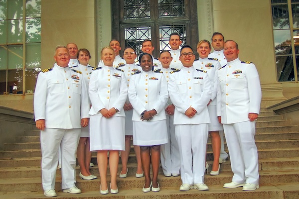 Fourteen graduates wear white navy uniforms on stone steps in front of a large building with ornate glass-paneled doors and columns.