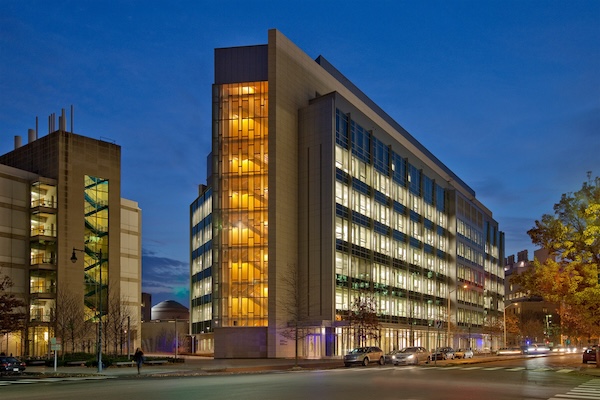 Modern glass building at dusk with warm orange-lit stairwell and bright interior lights along the street.