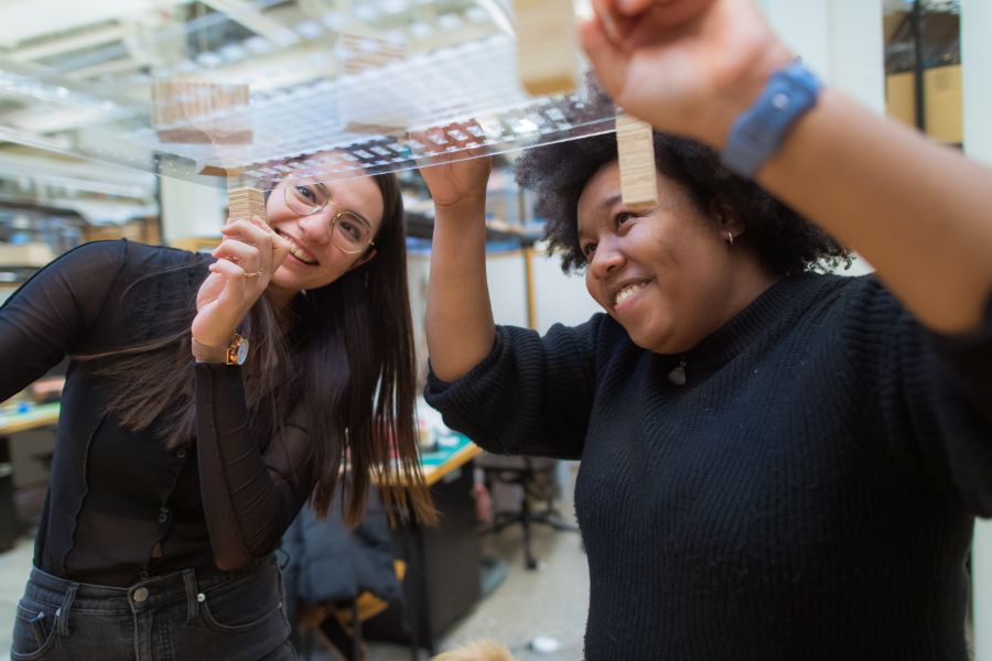 Two students smile as they work together on a transparent panel with components attached in a workshop or lab setting.
