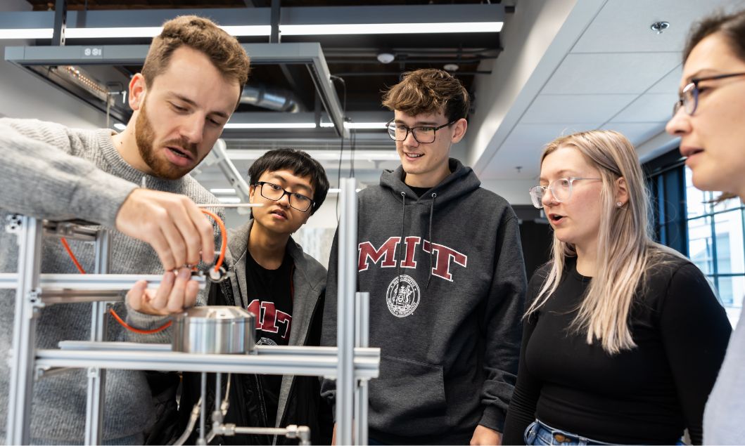 A group of students, some wearing MIT sweatshirts, gather around and examine a mechanical device in a modern classroom or lab setting.