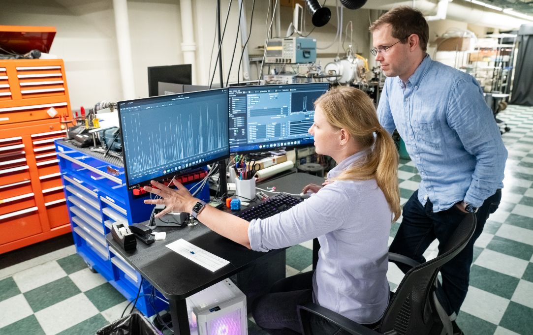 Two researchers review data on dual computer monitors displaying graphs in a lab filled with technical equipment and tool chests.