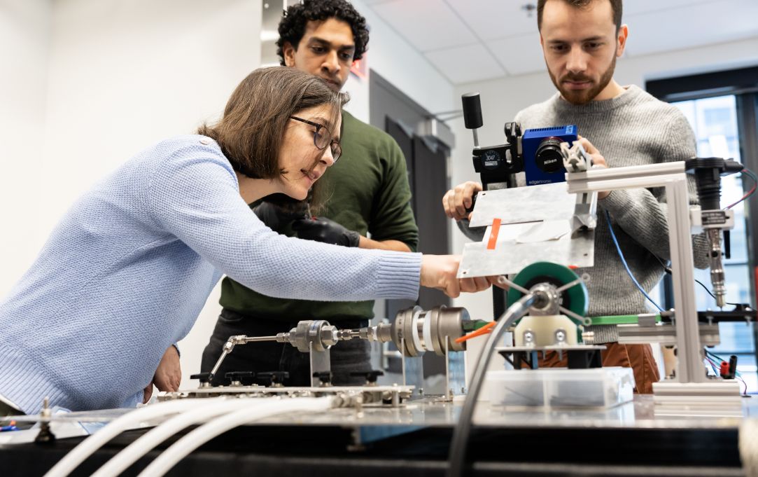 Three people closely examine and adjust a mechanical testing setup in a lab, with one person pointing at a component while another operates a camera.