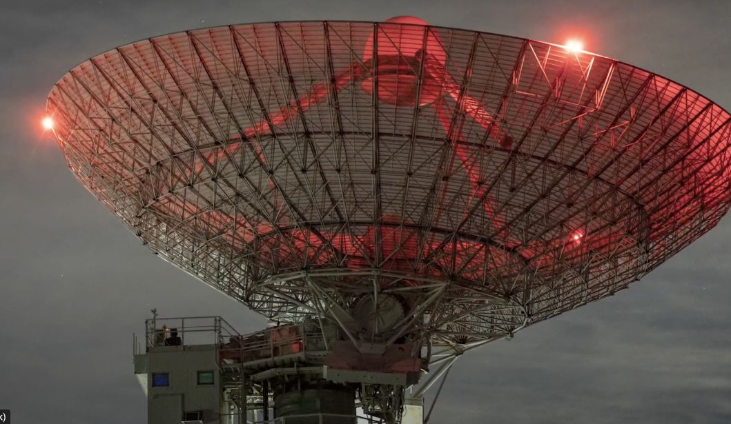 Large satellite dish glows with red lights against a cloudy night sky, mounted on a support structure.