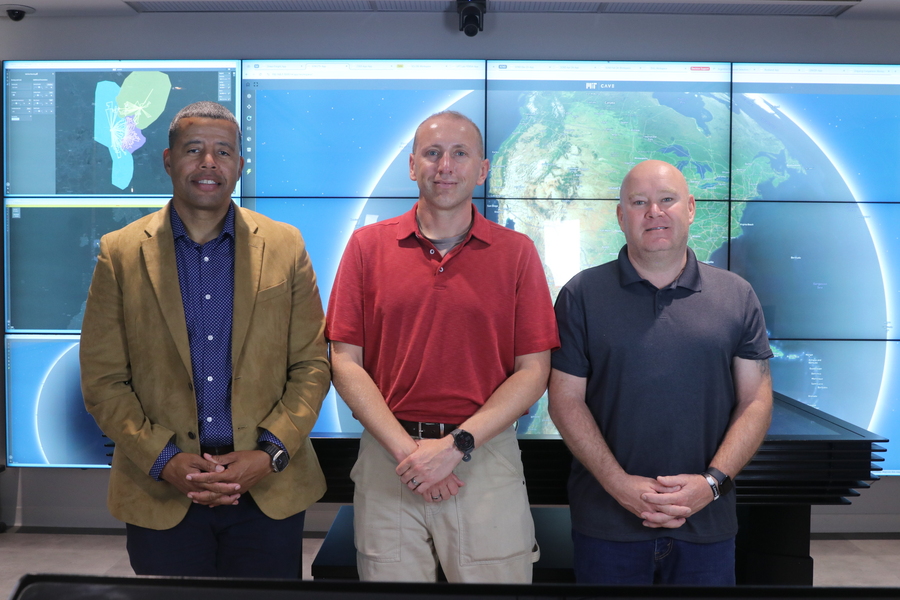 Three people standing in front of a large multi-screen display showing maps and satellite imagery.