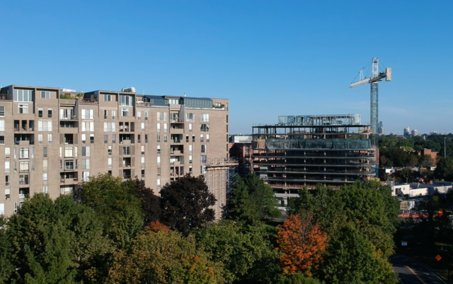 An existing apartment building stands beside a multi-story building under construction with a tower crane, surrounded by trees with early fall color.