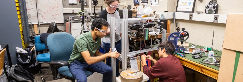 Three students wearing safety glasses collaborate on assembling a mechanical project in a cluttered workshop.
