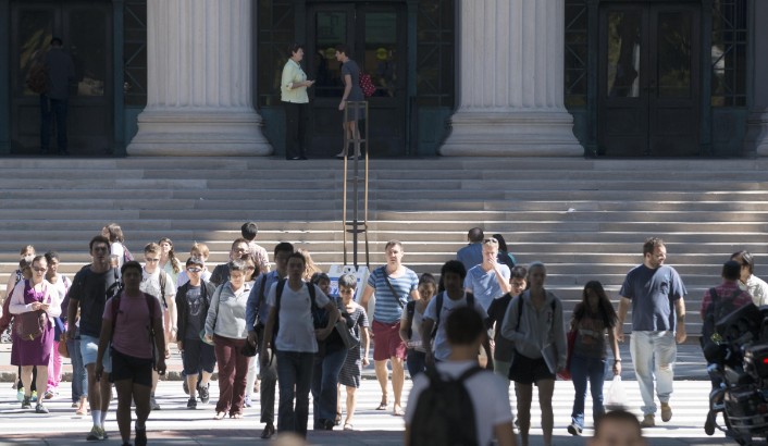 A large group of people walks away from MIT Building 7, which has tall columns and wide steps.