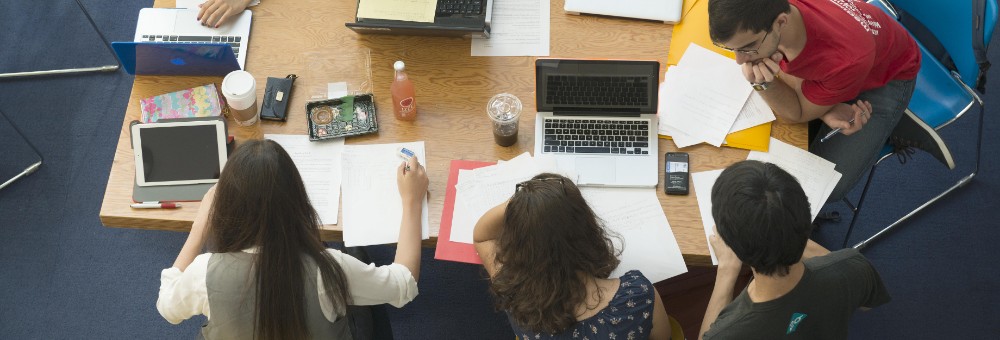 Overhead view of students working together at a table covered with laptops, papers, and drinks.