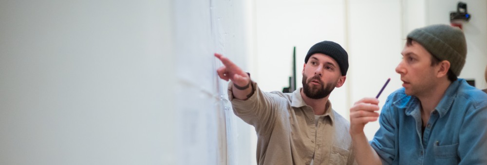 Two men in beanie hats discuss and point at a wall covered with large sheets of paper, possibly reviewing plans or designs.