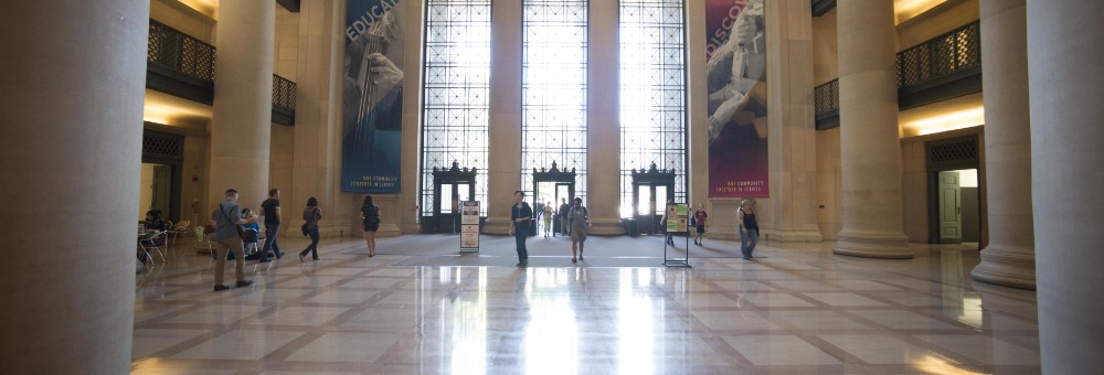 People walk through the grand lobby of MIT Building 7, with tall columns, large windows, and banners reading “Educate” and “Discover.”