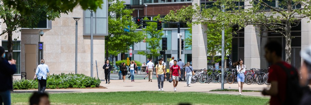 People walking through a green, sunlit campus area with bike racks and academic buildings in the background.