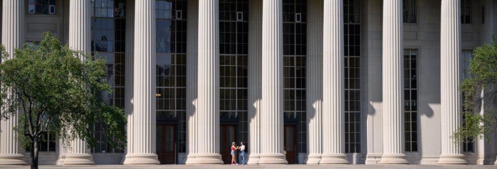 Two people stand in front of MIT Building 10 with tall columns and large windows, framed by trees.