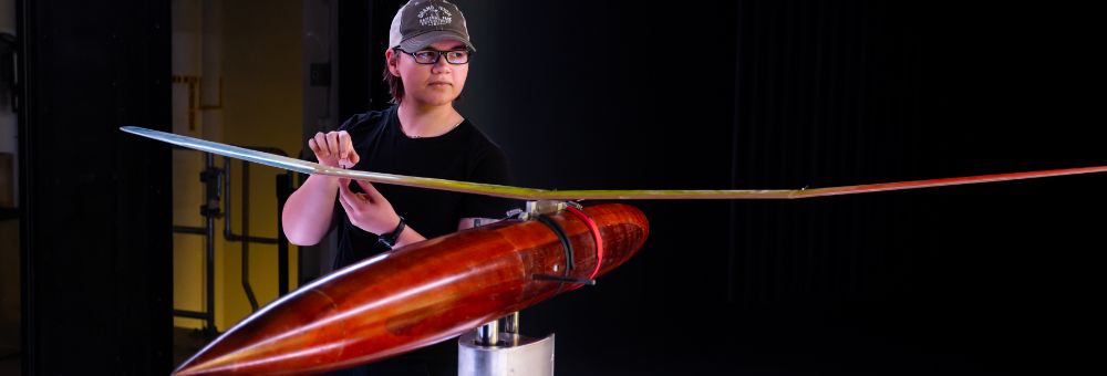 Student wearing glasses and a cap adjusts a sleek, aerodynamic model with long wings in a lab or testing space.