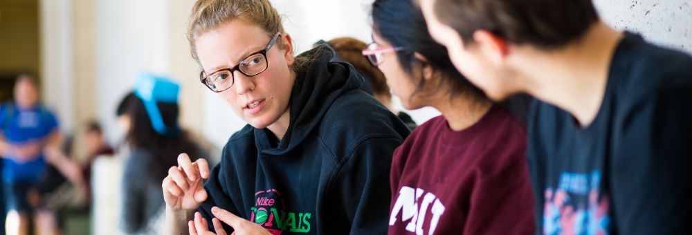 Three students sit in conversation, with one speaking and gesturing while the others listen attentively.