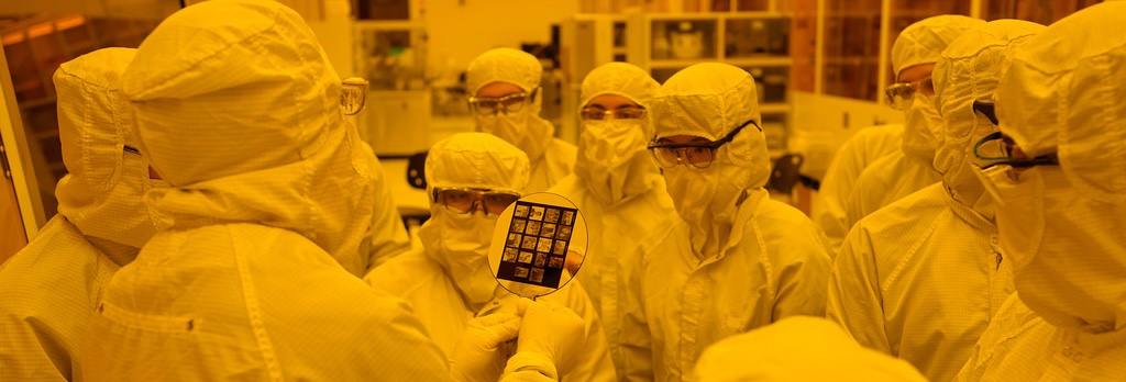A group of people in cleanroom suits gathers around a person holding a silicon wafer inside the MIT.nano building.