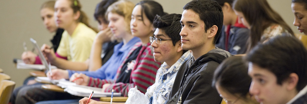 A group of students listens to a classroom lecture, with some taking notes.