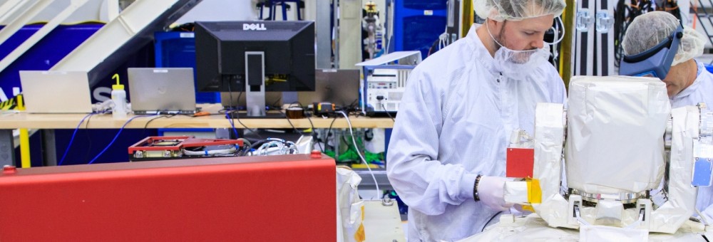 Two people in cleanroom suits work on a robotic device in a high-tech lab with computers and equipment in the background.